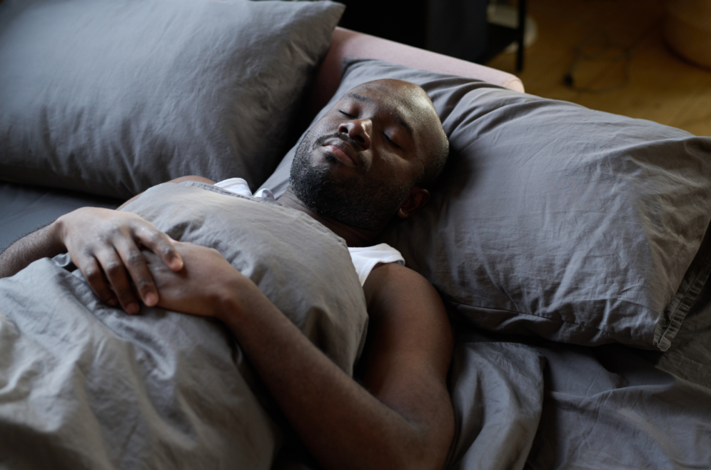 Man sleeping peacefully in his bed against a grey pillow with grey sheets. He has his hands resting across his chest. 