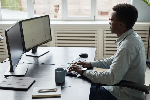 Young African American businessman sitting at his ergonomic workstation. He is typing on an external keyboard and looking at his two monitors that are about two feet away from him. 